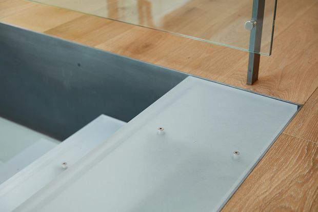 Close-up architectural detail of a stair opening or landing with pale timber flooring, a grey metal or concrete side wall, a light-colored stair or platform surface, and a frameless glass balustrade supported by slim metal posts. The image emphasizes construction materials and the transition between the floor and stair edge.