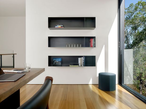 Bright, minimalist contemporary interior featuring a white wall with three recessed dark open shelving niches, a timber desk with a laptop and chair, and full-height corner glazing overlooking trees. Warm timber flooring and strong natural daylight create a clean home workspace atmosphere.