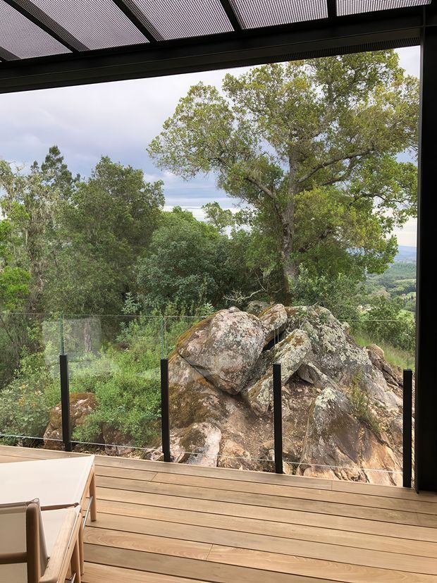A covered indoor-outdoor living space opens onto a timber deck with frameless glass balustrading, overlooking a large exposed rock outcrop, dense native vegetation, and a distant valley landscape. A slatted overhead roof provides shade, while part of a light timber dining table and chair is visible in the foreground. The scene has a strong connection to the natural, rocky site.