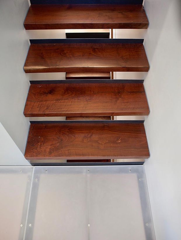 Close-up view looking up a compact stairway with thick, richly stained solid-wood treads cantilevered or fixed between pale walls. Dark gaps and visible structural elements separate the open risers, while the lower portion shows light metal or painted panel construction with exposed fasteners. The image focuses on the timber stair detailing and simple utilitarian construction.