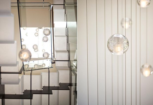 Interior architectural detail showing a contemporary stairwell with dark-edged glass balustrades and mirrored or highly reflective surfaces. A cluster of spherical pendant lights hangs through the stair void, while the adjacent wall is finished with vertical white panel lines. The composition emphasizes geometric repetition, transparency, and reflected light.