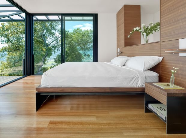 Bright contemporary bedroom with a low built-in timber platform bed, integrated bedside joinery, warm timber flooring and wall panelling, and large black-framed floor-to-ceiling glazing opening to leafy views. White bedding and a simple floral arrangement provide a clean, calm contrast to the warm wood surfaces.