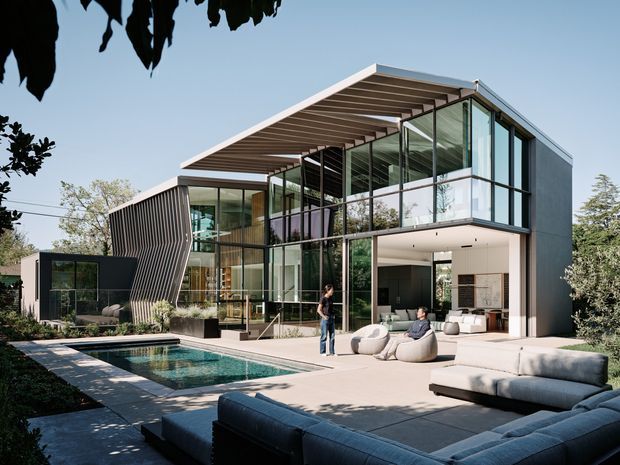Contemporary two-storey residence with extensive floor-to-ceiling glazing, a dramatic cantilevered upper-level roof canopy, and a dark solid side volume. The home opens onto a large paved outdoor entertaining area with a swimming pool, built-in and freestanding lounge seating, and landscaped planting. Strong indoor-outdoor connection is visible through the open ground-floor living area and transparent facade.