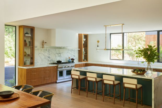 A bright, spacious open-plan kitchen with a large island and seating for four, dark green cabinetry, warm timber joinery, a veined marble backsplash, and integrated display shelving. The room features pale timber flooring, expansive black-framed windows, and a brass linear pendant light over the island. A dining table is partially visible in the foreground.