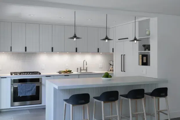 Bright, clean contemporary kitchen with pale vertical-panel cabinetry, a large island with seating for four, integrated appliances, a white tiled backsplash, and three black pendant lights. The palette is predominantly white and light grey with dark accents and light timber stool legs.