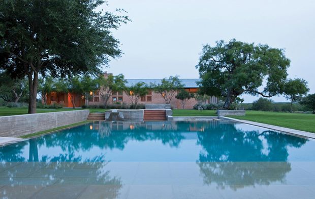 Wide exterior view of a low, horizontal brick residence set behind a large rectangular swimming pool. The pool reflects mature trees and the building, with broad paved surrounds, planted garden beds, lawns, and low masonry retaining or planter walls. The house has a warm brick facade, extensive glazing, and a shallow roofline, creating a calm, resort-like landscape setting.