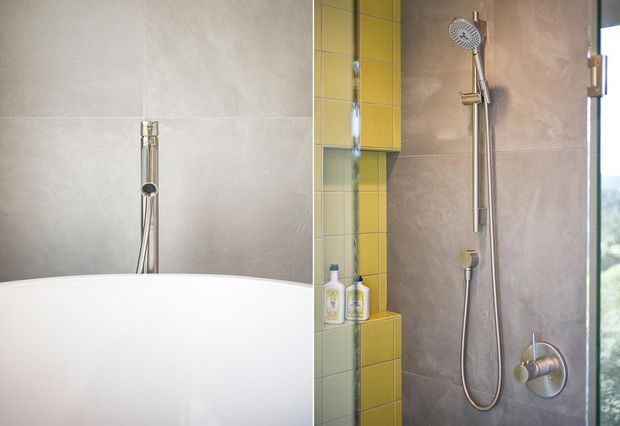 Close-up view of a contemporary bathroom shower and bathtub area. Large pale grey wall tiles surround a white freestanding bathtub, with a chrome bath filler or hand-shower fixture behind it. The adjacent shower has a chrome rail, handheld shower, round showerhead, and mixer controls, contrasted by a bright yellow tiled recessed shelf and vertical accent strip. Natural light enters through a window at the right.