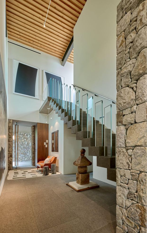 A spacious contemporary entry hall with a dramatic floating staircase and clear glass balustrade. The interior features a warm slatted timber ceiling, pale plaster walls, large-format neutral flooring, exposed natural stone walls, and timber doors. Sculptural wood decor and a small seating area sit beneath the stair, while a tall glazed opening brings daylight into the double-height space.