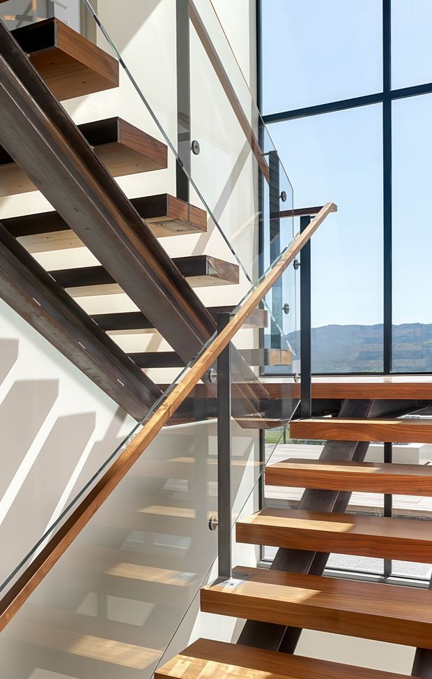 A bright contemporary stairwell with floating timber treads supported by dark steel stringers, clear glass balustrades, and timber handrails. Large floor-to-ceiling windows flood the stairway with natural light and provide views toward a hilly landscape. The open vertical composition emphasizes the stair structure and abundant glazing.