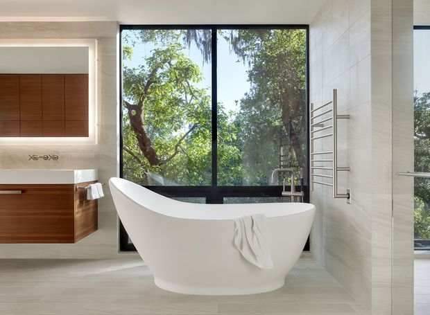 Bright contemporary bathroom featuring a sculptural freestanding white bathtub positioned in front of large black-framed windows overlooking leafy trees. The room has pale large-format tiled walls and floor, a timber floating vanity with white countertop, illuminated recessed mirror, wall-mounted fixtures, and a stainless towel warmer. A glass shower enclosure is visible at right.