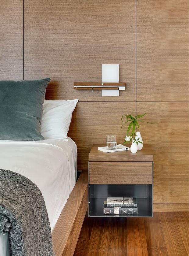 A close-up contemporary bedroom interior featuring a neatly made bed with white bedding and a dark grey-green cushion, a full-height warm timber wall panelled with fine horizontal grain and dark joint lines, and a compact timber bedside table with an open black storage compartment. A slim wall-mounted shelf and small white wall light sit above the table, while a small potted plant and books add restrained styling. The flooring is warm timber, with a textured grey throw in the foreground.
