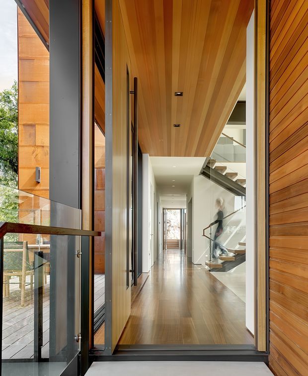 View through a glazed entry into a contemporary residential hallway. The entry corridor features extensive warm timber lining on the ceiling and walls, polished timber flooring, full-height glazing, dark metal-framed doors, and a sculptural open staircase with glass balustrades. The hallway extends toward a bright rear opening, creating a strong indoor-outdoor connection.