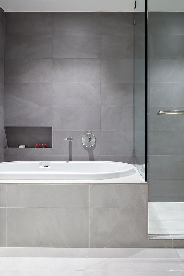 A contemporary bathroom featuring a built-in white bathtub set into a tiled surround, large-format grey wall and floor tiles, a recessed shelf with toiletries, and a frameless glass shower enclosure with chrome hardware.
