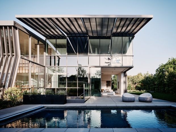 Contemporary multi-level residential house viewed from the pool, featuring extensive floor-to-ceiling glazing, a deep flat projecting roof canopy with exposed dark structural members, pale rendered or concrete surfaces, and landscaped garden surrounds. The reflective swimming pool and outdoor seating occupy the foreground, while the glass facade reveals interior living areas.