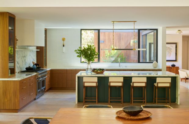 A warm, spacious contemporary kitchen with a large central island, integrated seating for four, timber cabinetry, dark green island joinery, and a pale stone benchtop. The room includes a marble-look splashback and fireplace surround at left, broad black-framed glazing overlooking a timber-clad exterior wall, pendant lighting, and an adjacent dining area visible at right. Natural timber and soft neutral finishes create a refined, inviting atmosphere.