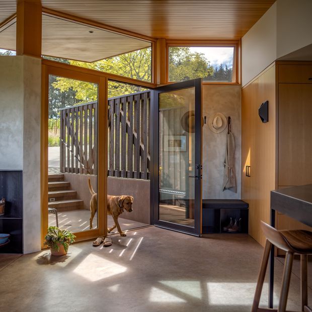 A warm, light-filled residential entryway with timber-framed glazing and a partially glazed timber-lined ceiling. The space opens to exterior steps and a fenced garden area, with a concrete floor, built-in timber storage, a dark entry door, bench seating, and a compact desk or console with chair at right. Natural daylight, timber detailing, and indoor-outdoor connection dominate the scene.