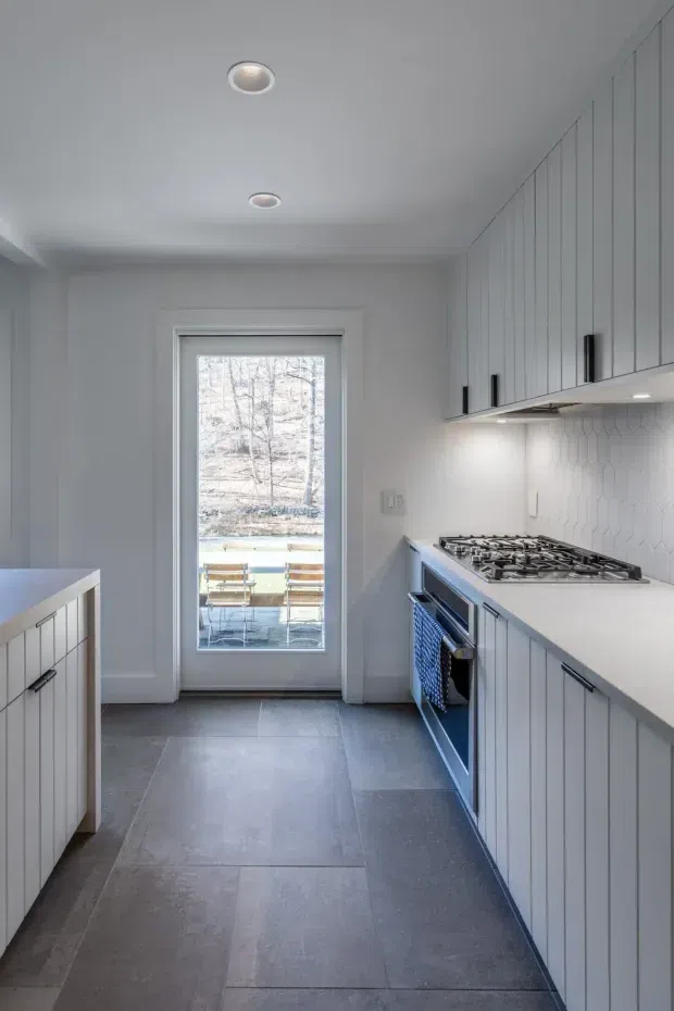 A narrow, bright galley kitchen with full-height pale grey shaker-style cabinetry, dark linear hardware, a long white benchtop, and a white geometric tile splashback. A gas cooktop and built-in oven sit along the right-hand run, while a compact island or peninsula is visible on the left. Large dark grey floor tiles lead to a glazed rear door opening onto a small outdoor deck. Recessed ceiling lights and under-cabinet lighting provide even illumination.
