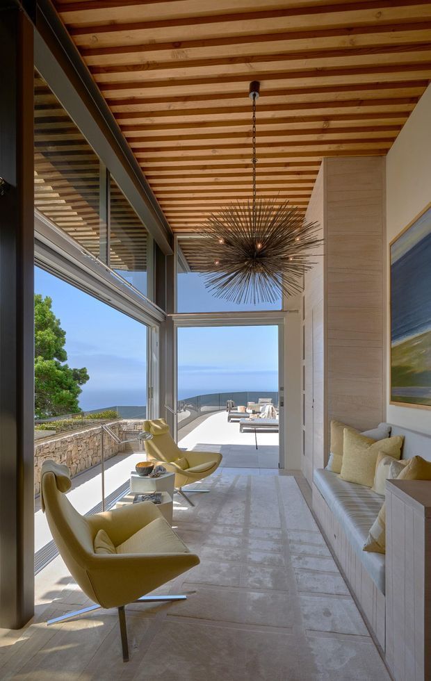 A bright, narrow coastal living space with full-height glazing opening to a terrace and expansive ocean views. The room features pale stone or concrete-look flooring, a built-in upholstered bench along one wall, sculptural mustard-yellow lounge chairs, a large spiky pendant light, timber slatted ceiling with exposed beams, dark metal window framing, and light vertical wall panelling. A stone retaining wall and outdoor seating are visible beyond the glazing.