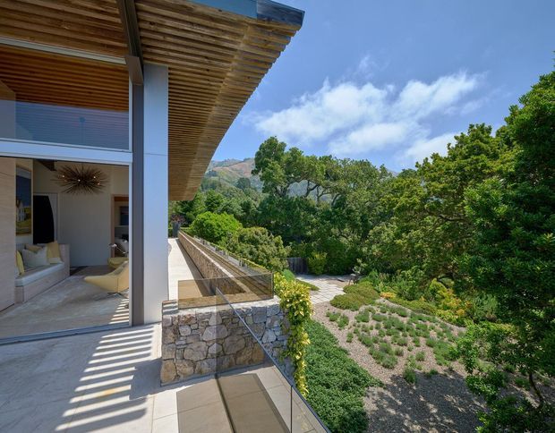 Exterior view of a modern home opening onto a landscaped terrace and garden. The image highlights a deep timber slatted roof overhang, full-height glazed sliding doors, pale structural columns, stone retaining walls, and a transparent glass balustrade. A contemporary living room is visible through the glazing, with a mountainous, tree-filled setting beyond.