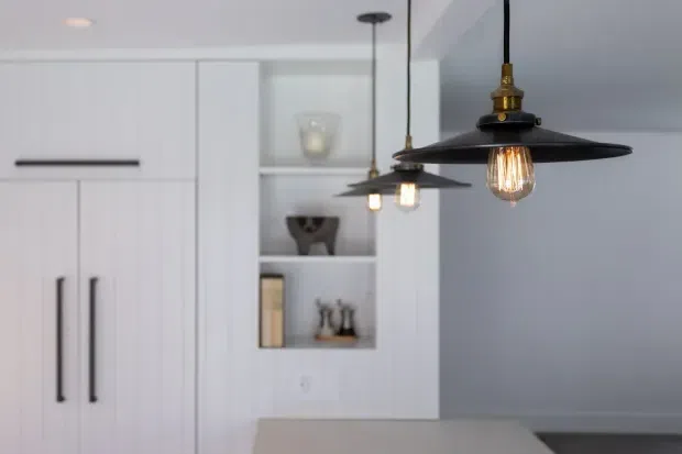 Close-up view of a refined white kitchen with tall flat-panel cabinetry, long black bar handles, open display shelving, and a pale benchtop. Three black pendant lights with exposed warm filament bulbs hang above the island, creating a contemporary industrial accent against the otherwise clean, light-toned interior.