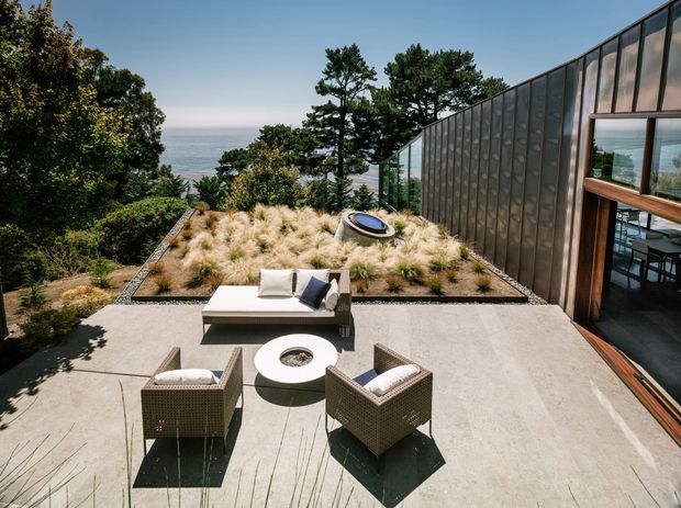 A modern coastal home viewed from an elevated outdoor terrace. The expansive concrete-paved patio contains woven lounge chairs, a low fire-pit table, and a built-in white bench with cushions. Beyond it is a landscaped rooftop or terrace garden with ornamental grasses, shrubs, and a circular skylight or roof feature. A tall glazed and dark-framed building facade opens onto the terrace, with ocean views visible through the trees.