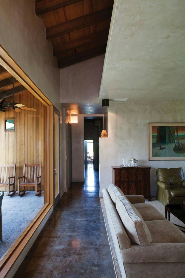 A tall, warm-toned living room interior with a high sloped timber ceiling and exposed dark wood beams. A large timber-framed glazed opening connects the room to a covered outdoor seating area. The space has polished dark flooring, textured plaster walls, a neutral sectional sofa, leather armchair, timber sideboard, pendant lighting and framed artwork. A hallway recedes toward the rear, creating a strong axial view.