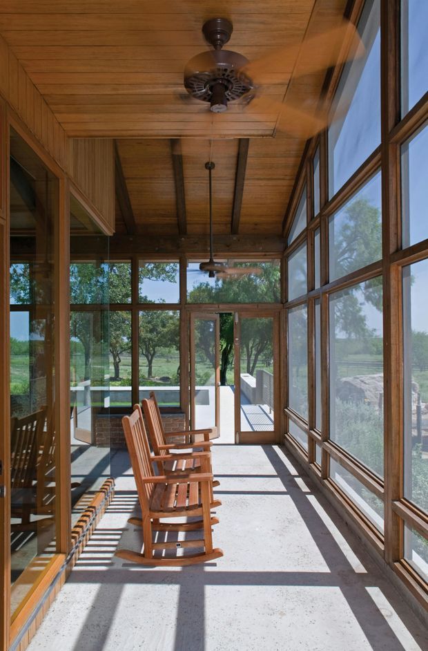 A long, bright enclosed sunroom or screened porch with extensive timber-framed glazing on both sides, a warm timber-plank ceiling with exposed beams, and a pale concrete floor. Several wooden rocking chairs line the space, which opens onto an outdoor terrace and landscaped setting. Ceiling fans provide ventilation, while strong sunlight creates long geometric shadows across the floor.