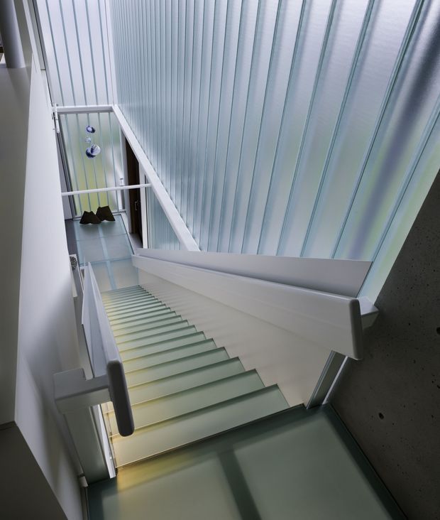 A modern interior stairway viewed from above, with a long flight of pale stairs, translucent glass or frosted-glass treads and balustrade elements, white angular handrails, and a tall ribbed translucent wall or ceiling creating a bright vertical atrium. A dark exposed concrete wall contrasts with the predominantly white and cool grey palette. Pendant lights are visible at the lower landing.