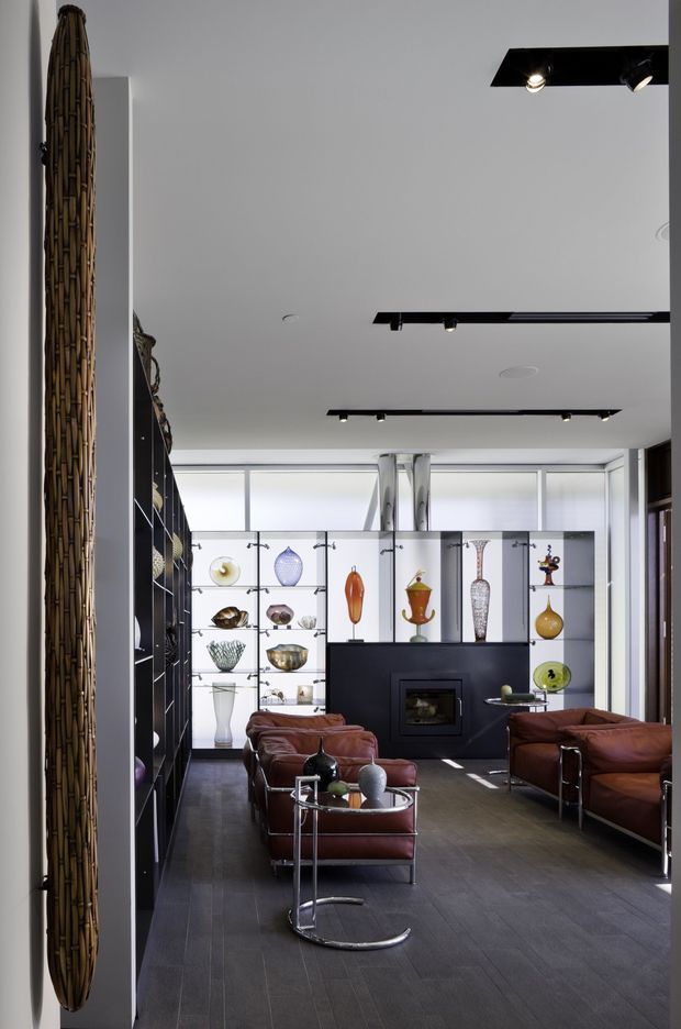 A sophisticated modern living room with a dark tiled floor, white walls, black built-in shelving, and a dramatic black fireplace feature wall. The room is furnished with brown leather sofas and chrome-framed lounge chairs, while a backlit display wall showcases decorative glass and ceramic objects. Linear black track lighting and large clerestory-style windows contribute to the contemporary gallery-like atmosphere.
