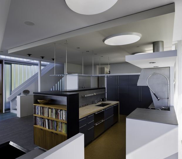 A contemporary open-plan kitchen viewed from an adjacent living or circulation area. The space features dark blue-black cabinetry, a timber breakfast-bar or shelving unit filled with books, dark flooring, pale benchtops, integrated appliances, a sculptural curved extractor hood, pendant lights, and large glazed openings beside a stair. Circular ceiling lights and crisp white surfaces create a clean, modern interior.