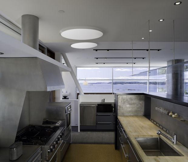 A contemporary galley-style kitchen with dark charcoal cabinetry, stainless-steel appliances, concrete-toned surfaces, and a long timber benchtop with an integrated double sink. Large floor-to-ceiling windows open to a bright waterfront or coastal outlook, while sculptural circular ceiling lights and exposed pendant track lighting add architectural interest.