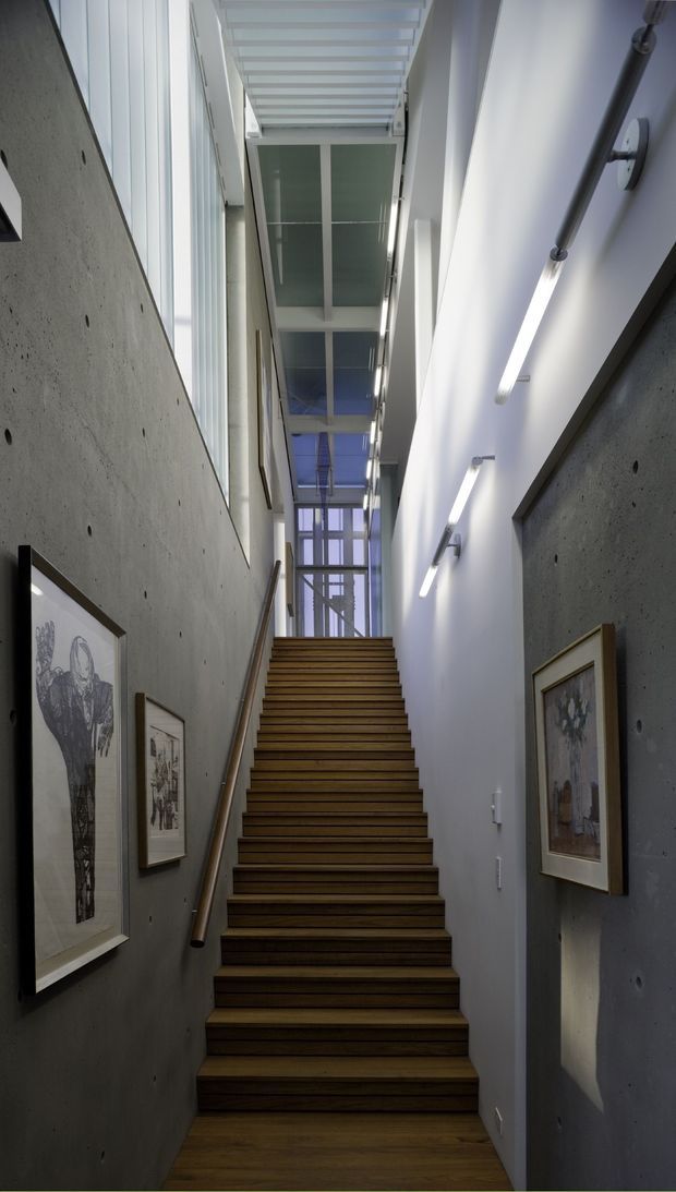 A narrow, tall stairway with dark timber treads rises between exposed grey concrete walls and smooth white walls. Linear wall-mounted lights illuminate framed artwork, while a glazed roof and clerestory-like overhead glazing bring daylight into the vertical circulation space. The design has a refined contemporary, gallery-like character with strong verticality and contrasting concrete, timber, glass, and white surfaces.