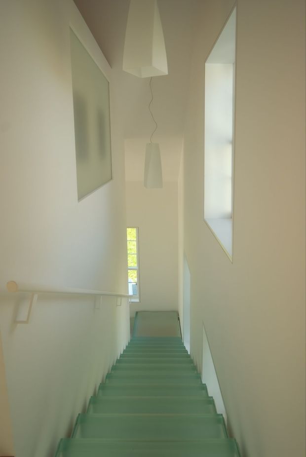 A minimalist interior stairway viewed looking down a narrow flight of translucent green-tinted glass steps. The stairwell has smooth off-white walls, a simple pale handrail, tall narrow glazing at the landing, and sculptural white pendant lights suspended in the double-height void. The space is bright, calm, and highly geometric.