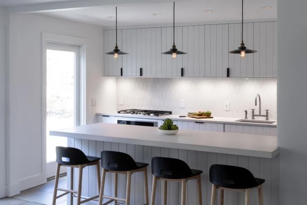 Bright, refined contemporary kitchen with a large pale island and four black bar stools. The kitchen features white vertical-panel cabinetry, a light hexagonal tile backsplash, integrated appliances, a stone-look benchtop, and three dark pendant lights. A glazed exterior door provides natural light.
