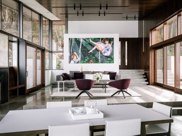 A sophisticated open-plan interior combining a dining area in the foreground with a spacious living room beyond. Floor-to-ceiling glazing and timber-framed windows surround the room, bringing in views of trees. The living area features contemporary lounge seating, a low coffee table, a large artwork, and a short flight of steps leading upward. A dark timber ceiling and wall paneling contrast with white walls, pale stone flooring, and streamlined furniture.