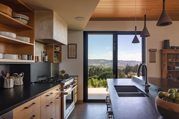 Warm, contemporary kitchen with timber cabinetry, open shelving, dark benchtops, stainless steel appliances, and a large island with an integrated double sink. Full-height glazing and a glazed door open to a landscaped outlook and distant hills. The ceiling combines a timber slatted section with painted plasterboard, and three dark pendant lights hang above the island.