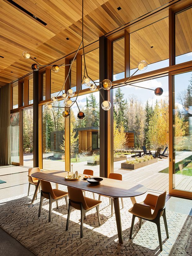 A warm, light-filled dining area with a long timber dining table and contemporary upholstered dining chairs. The room features extensive floor-to-ceiling glazing with dark timber frames, a richly timber-lined ceiling, and a sculptural multi-globe pendant light. Through the glazing, a landscaped outdoor deck and wooded garden are visible.
