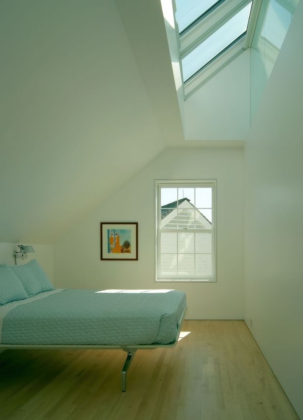 A bright, minimalist bedroom with a suspended or platform-style bed, pale turquoise quilt and pillows, light timber flooring, white walls, and a small framed artwork. A large high-level skylight brings in abundant natural light, while a multi-pane window overlooks a neighbouring roof. The room has a clean, airy appearance with minimal furnishings.