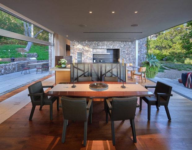 Open-plan dining and kitchen space with floor-to-ceiling glazing opening onto landscaped outdoor areas. A large timber dining table with dark upholstered chairs occupies the foreground, while a stone-clad kitchen wall, island, exposed shelving, and integrated appliances form the backdrop. Warm timber flooring contrasts with the cool grey stone and dark ceiling, creating a refined indoor-outdoor setting.