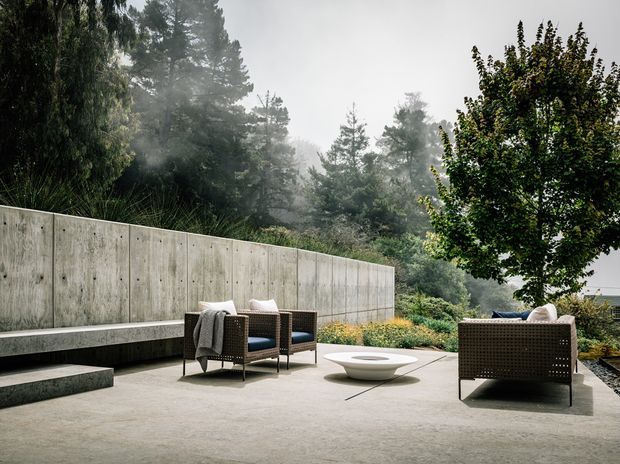 A refined outdoor terrace set within a misty, wooded landscape. The space features a broad smooth concrete-paved surface, a tall exposed concrete retaining wall with an integrated linear bench, dark woven outdoor armchairs and sofa, and a low sculptural fire bowl or coffee table. Dense trees and layered planting surround the terrace, creating a secluded, atmospheric setting.