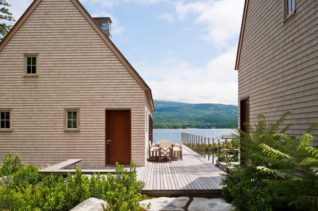 Exterior view of two compact shingle-clad buildings framing a timber deck and walkway leading toward a calm lake, with forested hills beyond. The left building has a steep gable roof, small windows, and a dark timber entry door; the right building has matching shingle cladding. Native-style planting, rocks, outdoor dining furniture, and a simple metal balustrade complete the lakeside setting.