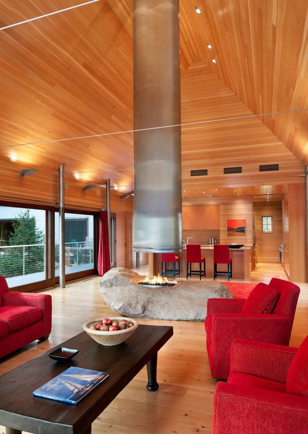 A dramatic open-plan living room with a very high, timber-lined vaulted ceiling and warm timber wall panelling. The room features expansive glazing, multiple red upholstered lounge chairs, a dark timber coffee table, and a sculptural stone fireplace with a suspended cylindrical metal flue and open fire. A compact kitchen and dining area are visible beyond the living space. Recessed ceiling lights and linear ventilation grilles are integrated into the timber ceiling.