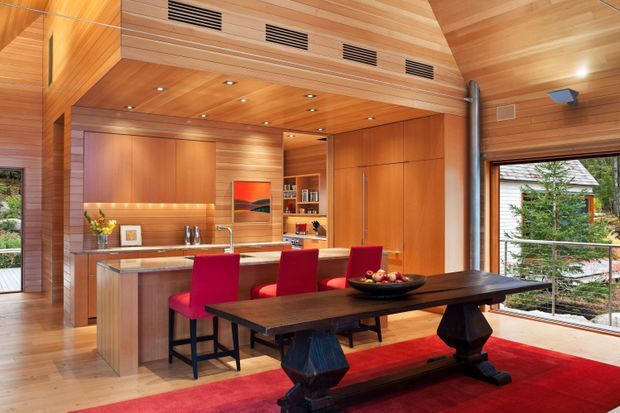 Open-plan kitchen and dining area clad extensively in warm timber, with a vaulted timber ceiling, exposed ventilation grilles, recessed lighting, and large windows. The kitchen has integrated timber cabinetry and a pale stone benchtop with a central island and red bar stools. A long dark timber dining table with bench seating sits on a bold red rug, creating a warm contemporary interior.