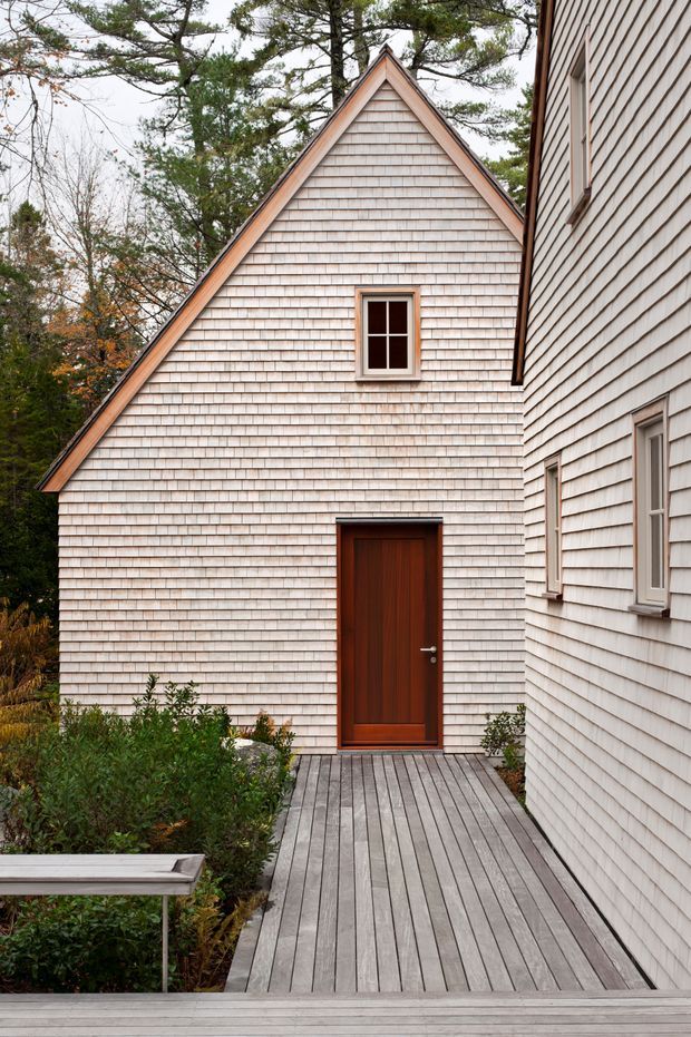 Exterior view of a small gabled building connected to a larger shingle-clad structure. The facade is covered in pale wood shingles, with a steep triangular gable, a small divided window, and a dark timber entry door. A weathered grey timber deck and walkway lead through landscaped planting, while mature trees surround the building.