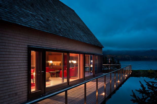 Exterior evening view of a waterfront house with a steep dark shingle roof, shingle-clad walls, extensive floor-to-ceiling glazing, and a timber deck extending toward the water. Warm interior lighting and integrated deck lights contrast with the cool blue dusk setting.