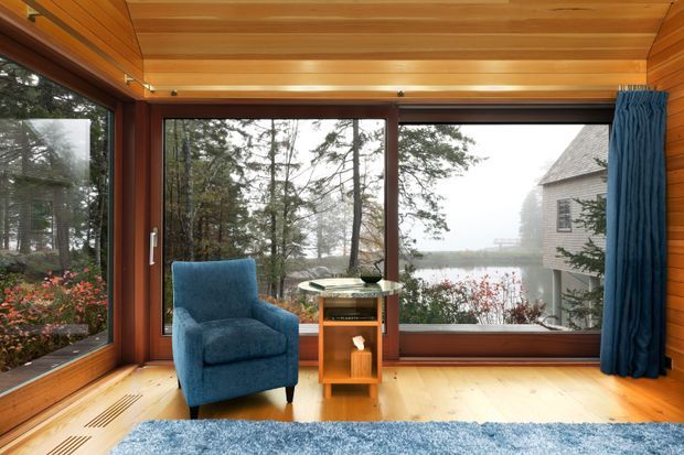 A compact wood-lined sunroom or enclosed living space with extensive corner glazing overlooking a wooded, misty landscape and water. Warm timber wall and ceiling panelling, timber-framed windows, pale timber flooring, a blue upholstered armchair, a small timber side table, blue curtains, and a blue rug create a calm retreat-like interior.