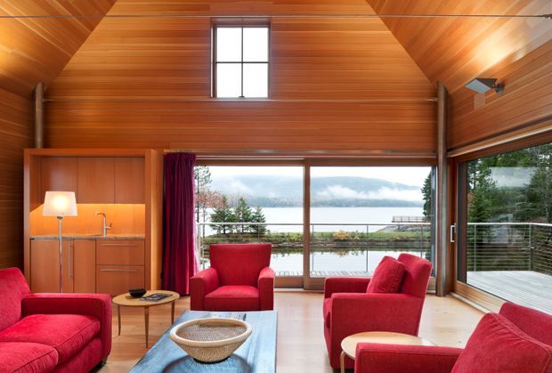 Warm, timber-lined living room with a steeply pitched, vaulted ceiling and exposed structural posts. The room features large windows and sliding glazing opening to a deck, with views of a landscaped waterfront setting. Red upholstered lounge chairs and sofas surround a low coffee table, while a compact built-in timber kitchenette or bar joinery occupies the left wall.