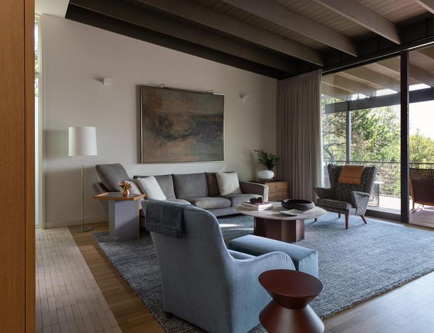 A refined contemporary living room with exposed dark structural beams and a high ceiling, large floor-to-ceiling glazing, and views to a leafy outdoor terrace. The room contains a grey sectional sofa, upholstered lounge chairs, a round timber coffee table, side tables, a floor lamp, a large area rug, curtains, and a neutral-toned feature artwork. Warm timber flooring and muted grey furnishings create a calm, sophisticated atmosphere.
