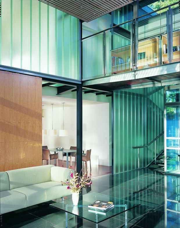 A sophisticated double-height modern interior featuring a glazed mezzanine or upper-level walkway, exposed dark steel framing, translucent green ribbed wall panels, and a slatted timber ceiling detail. The lower level combines a living room with a pale upholstered sofa, glass coffee table, and floral arrangement, opening toward a dining area with a dark table and pendant lights. Reflective dark flooring, extensive glazing, and a stairway reinforce the dramatic, light-filled architectural character.