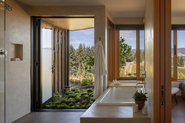 Warm contemporary bathroom with a freestanding rectangular bathtub set into a stone-look surround, a wall-mounted shower, and large timber-framed glazing. An open glass door connects directly to a planted outdoor area, while a partially visible adjoining room appears beyond the timber sliding doors. Natural light, textured plaster walls, timber frames, and garden views create a calm indoor-outdoor atmosphere.